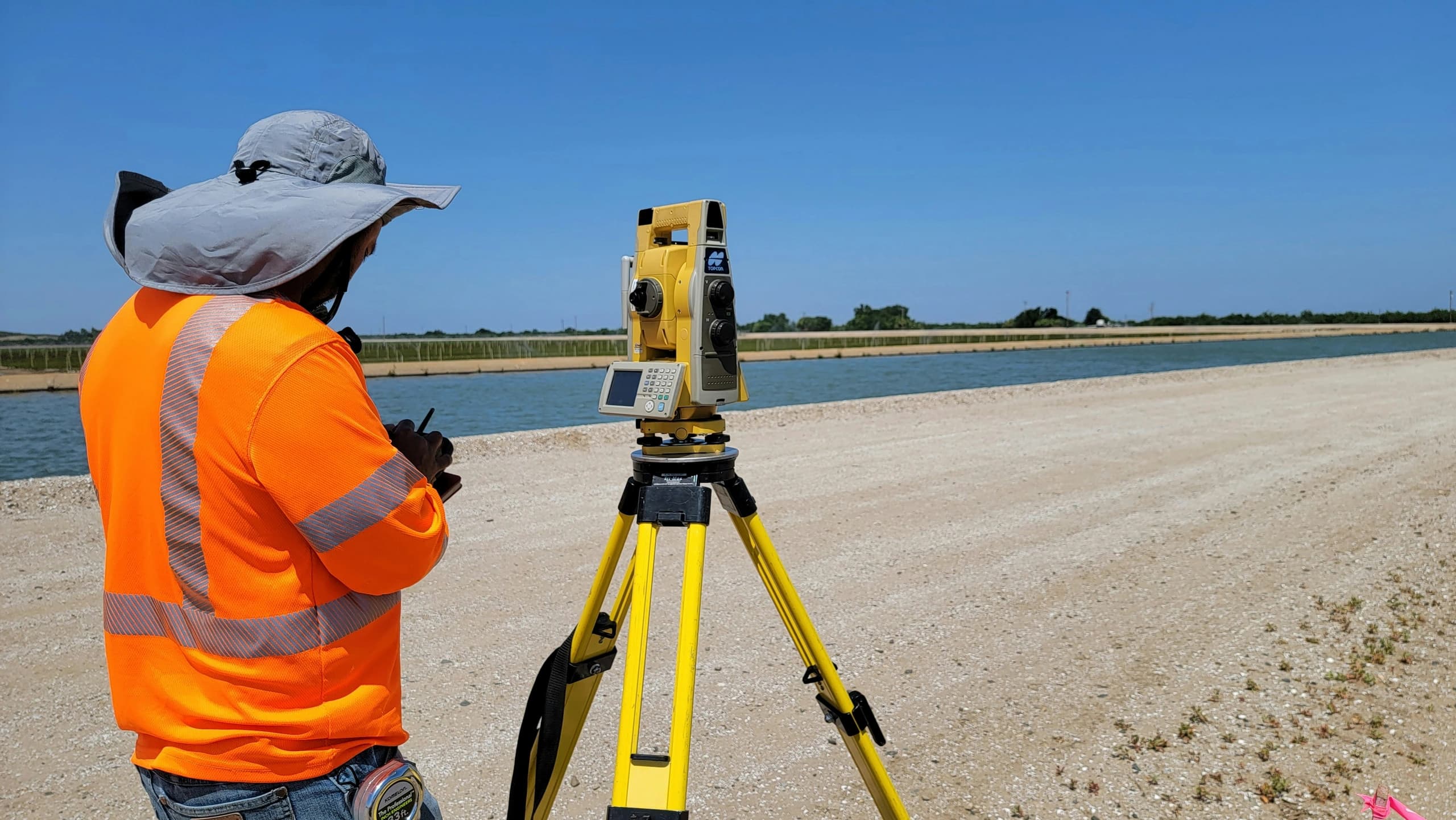 Surveyor in a high-visibility vest recording readings beside a tripod-mounted total station