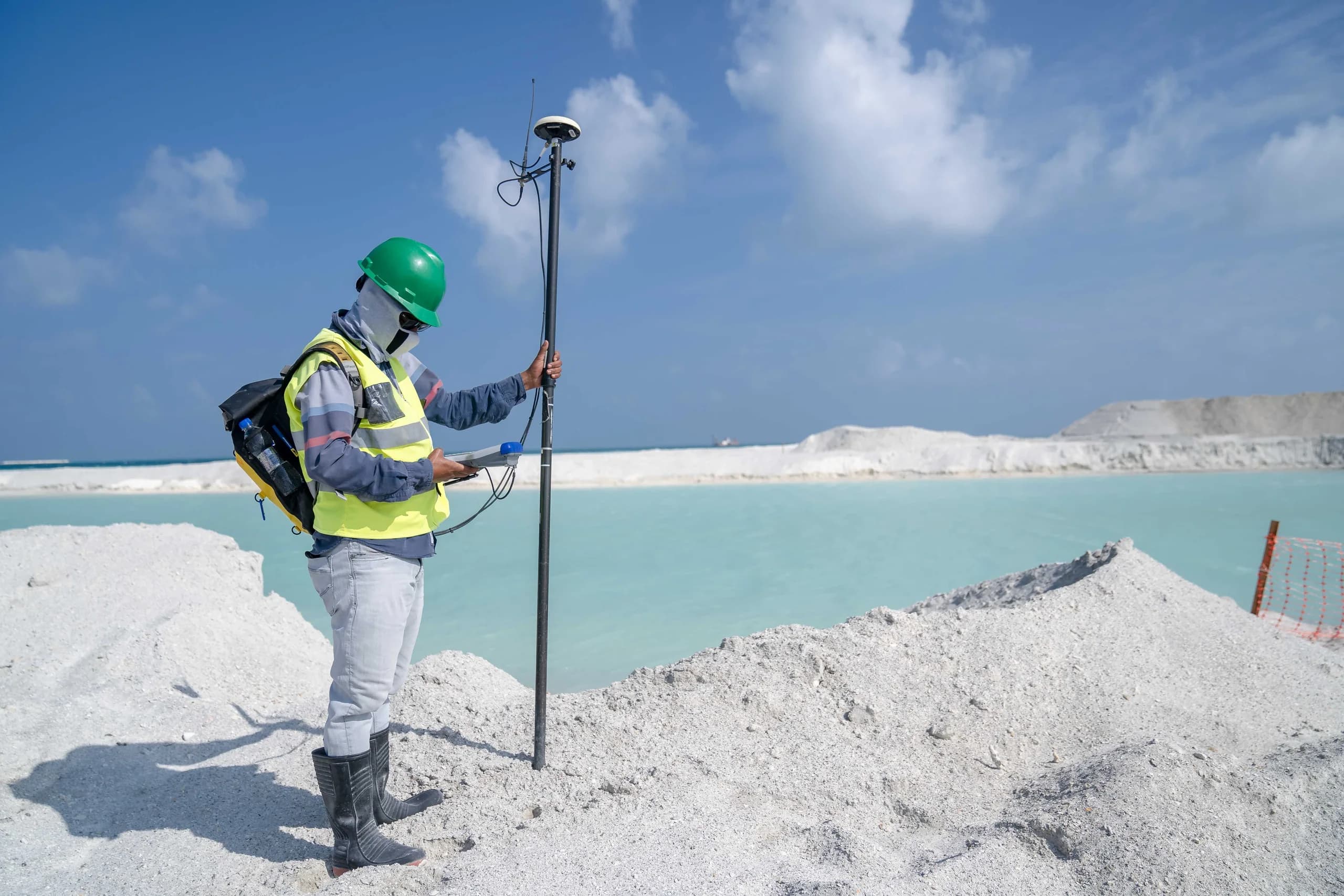 Land surveyor in a hard hat and high-visibility vest operating a GNSS receiver on a survey pole, standing on white sand fill beside turquoise water at a coastal reclamation site