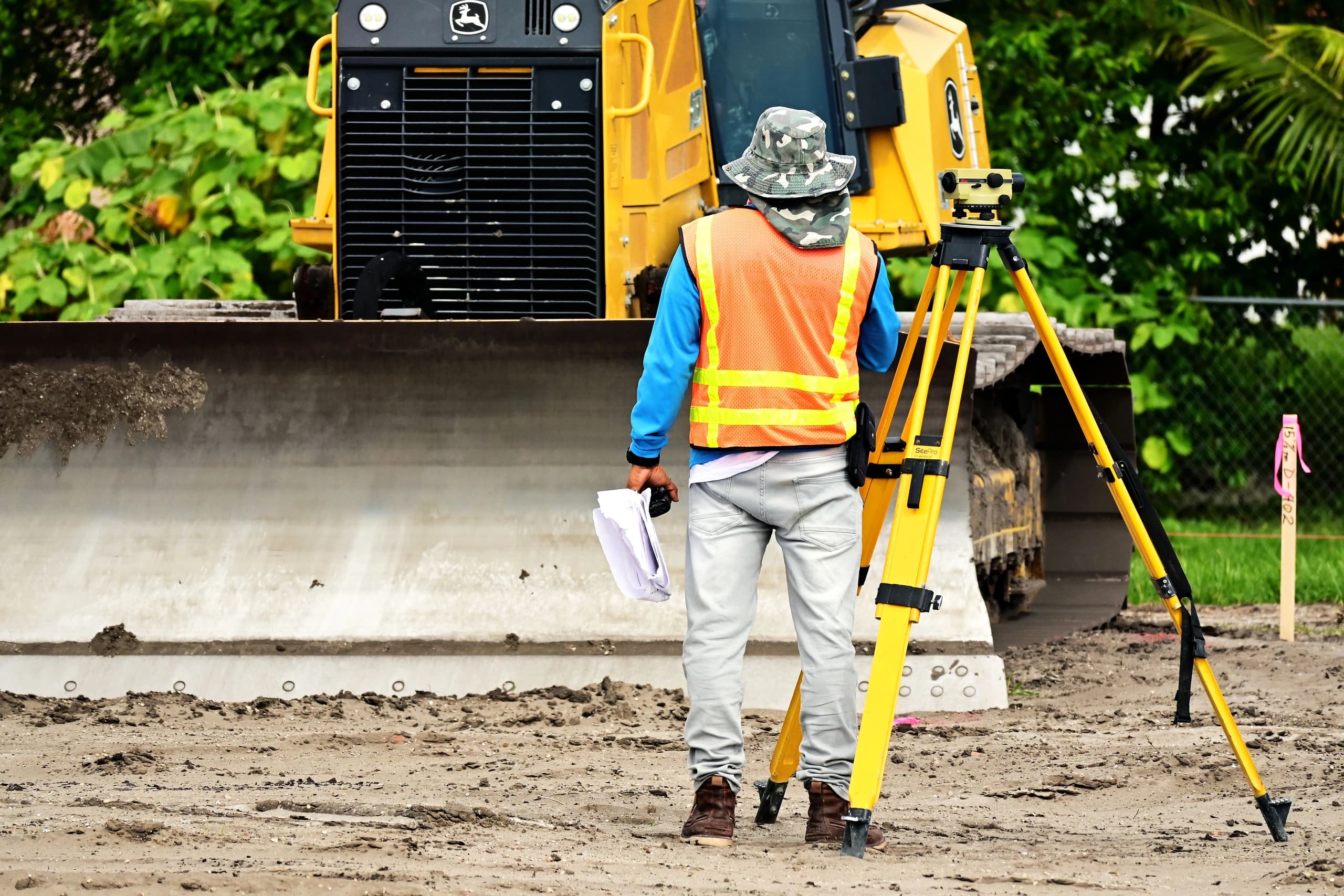 Surveyor in a high-visibility vest holding rolled drawings beside a tripod-mounted optical level on a construction site, a yellow bulldozer and dense green vegetation behind