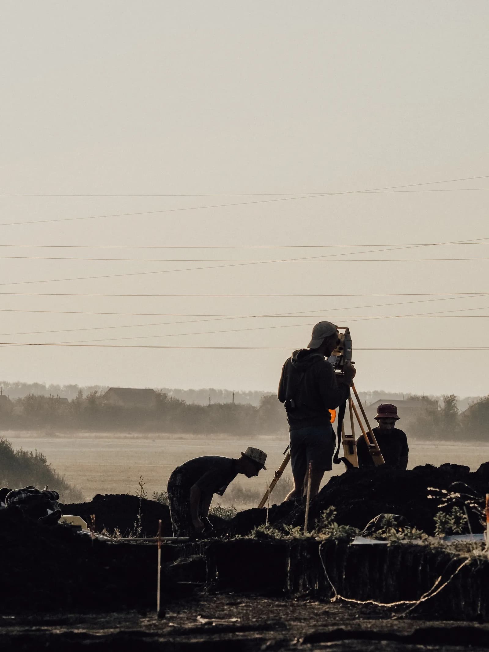 Surveyors silhouetted at dusk, one sighting through a total station on a tripod above freshly excavated ground